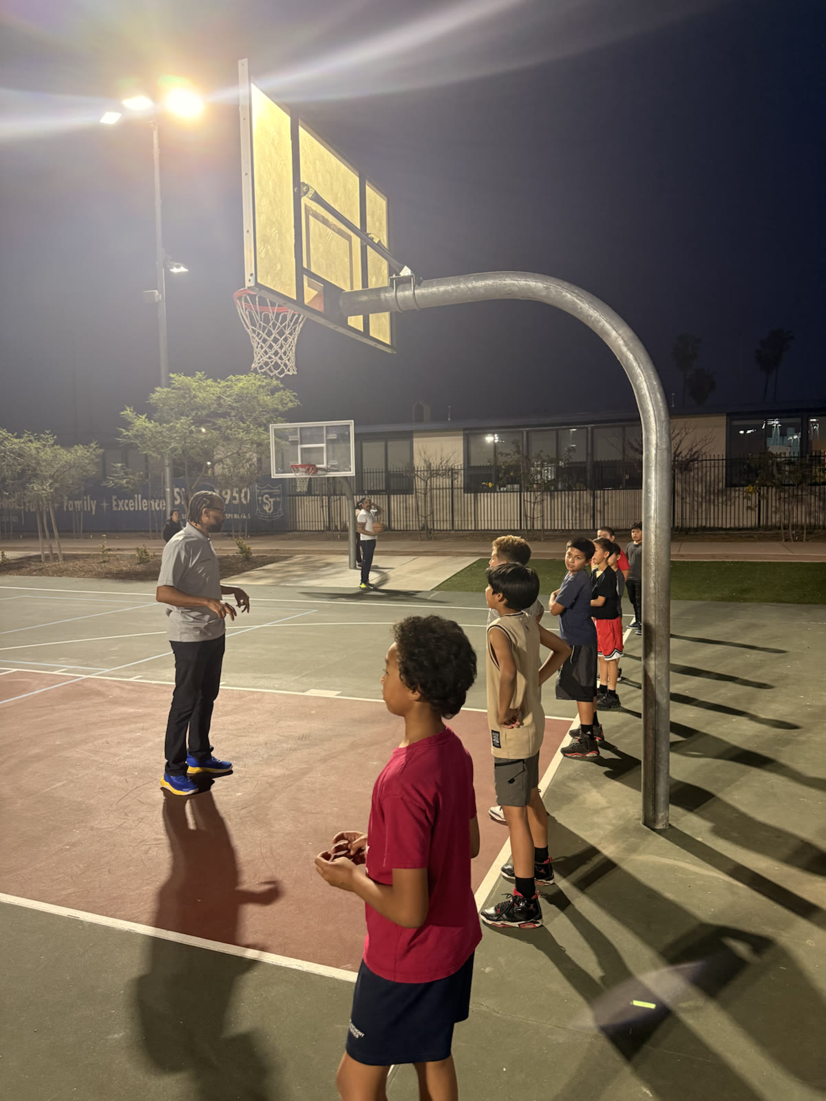 A coach speaking to young players lined up beneath an outdoor basketball hoop at night